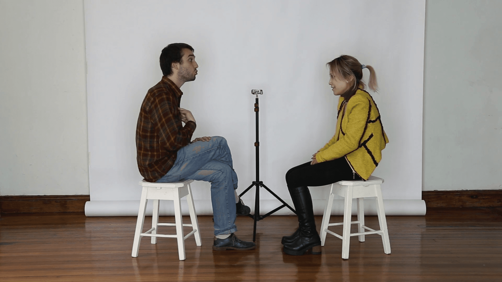 A man and woman sit on stools facing each other, with a microphone between them