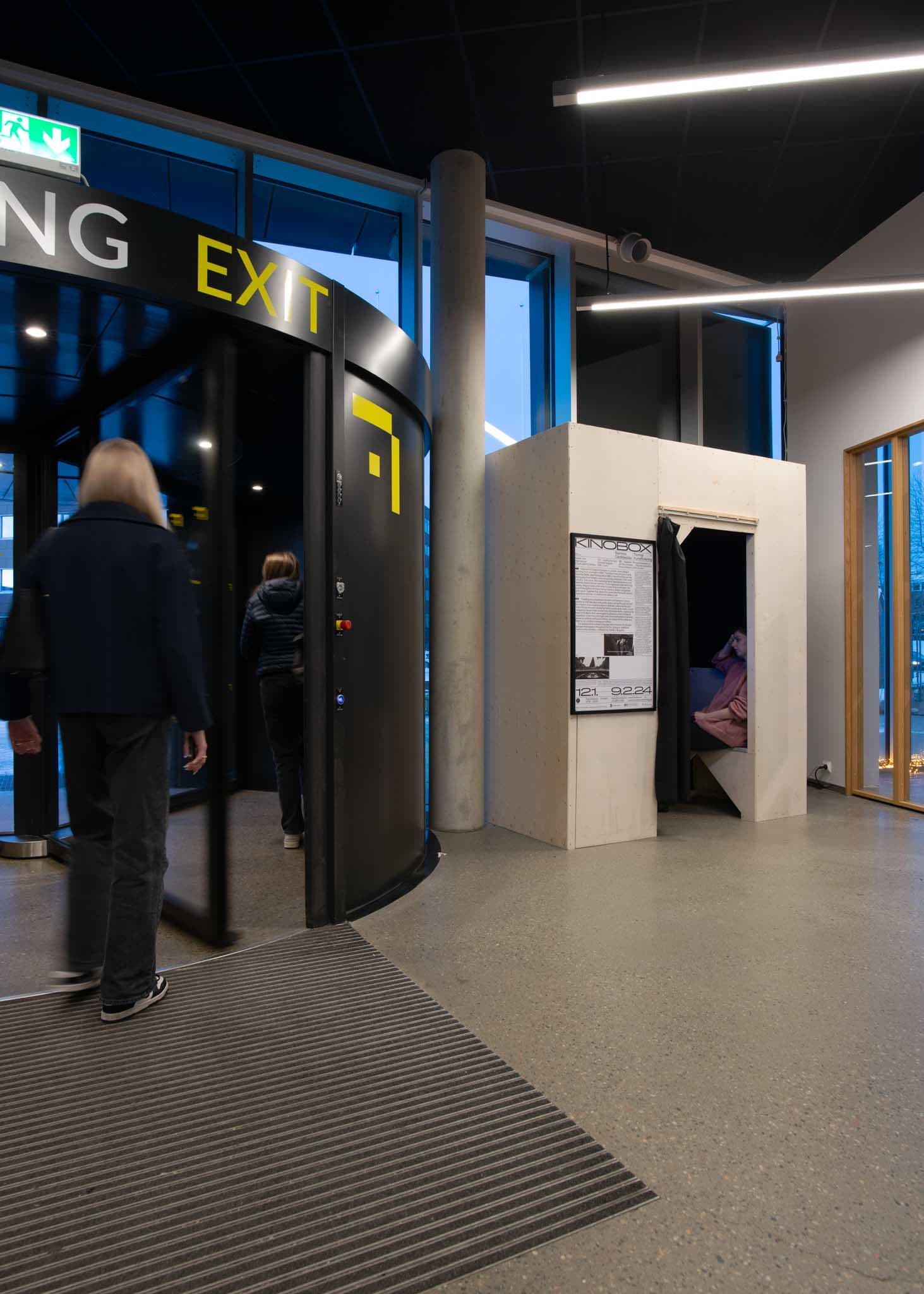 A small wooden box-cinema stands to the right of a black revolving door.