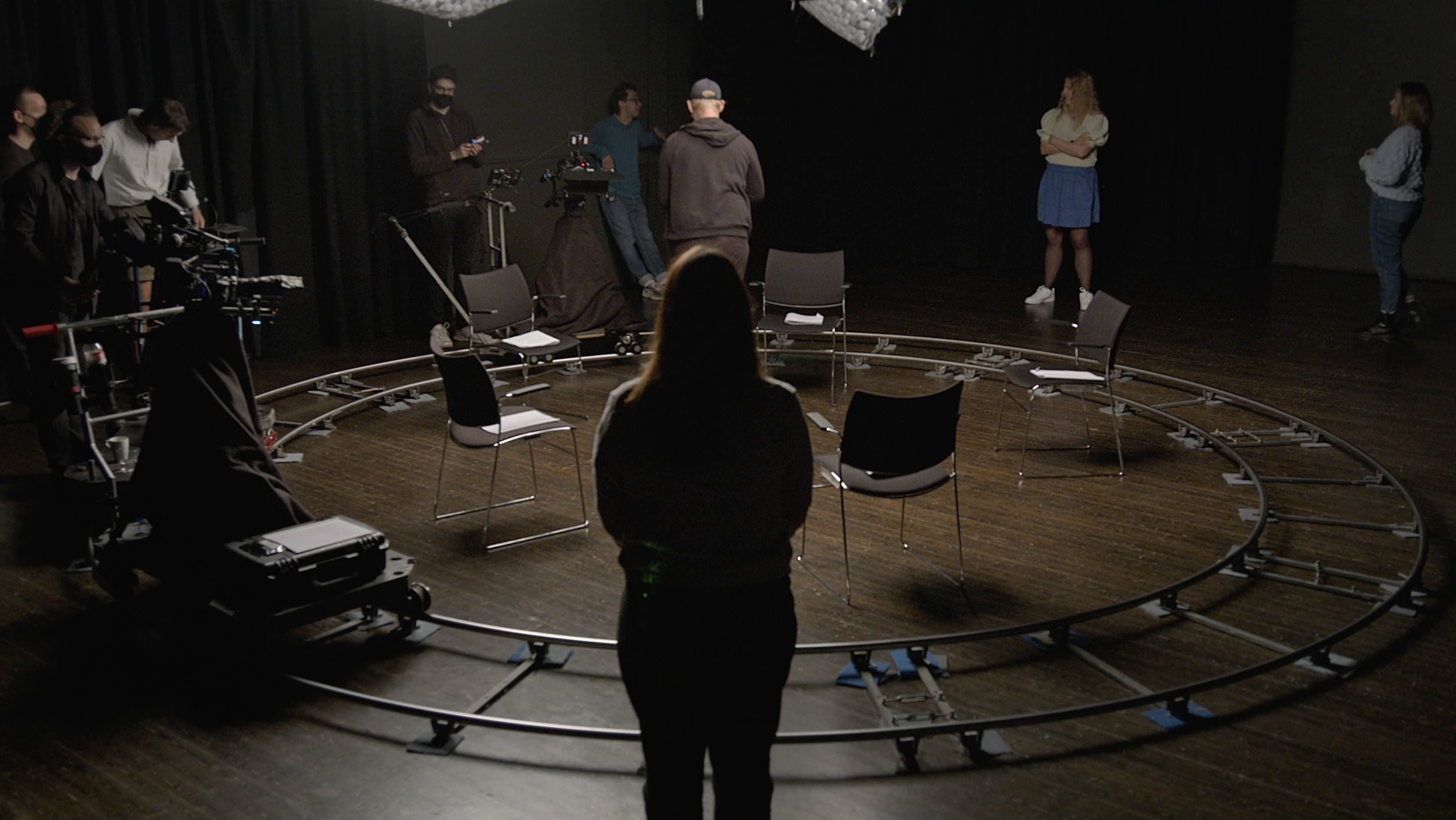 People stand around in a theatre blackbox. A camera track encircles 5 chairs set in a circle