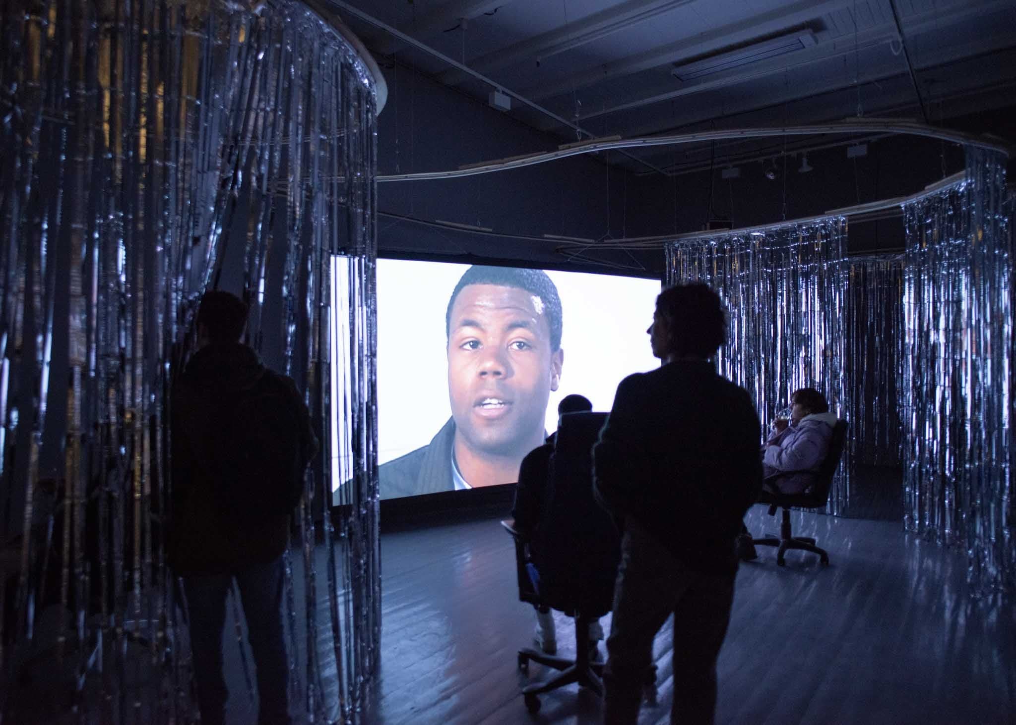 People sit in office chairs, watching a film in a dark gallery