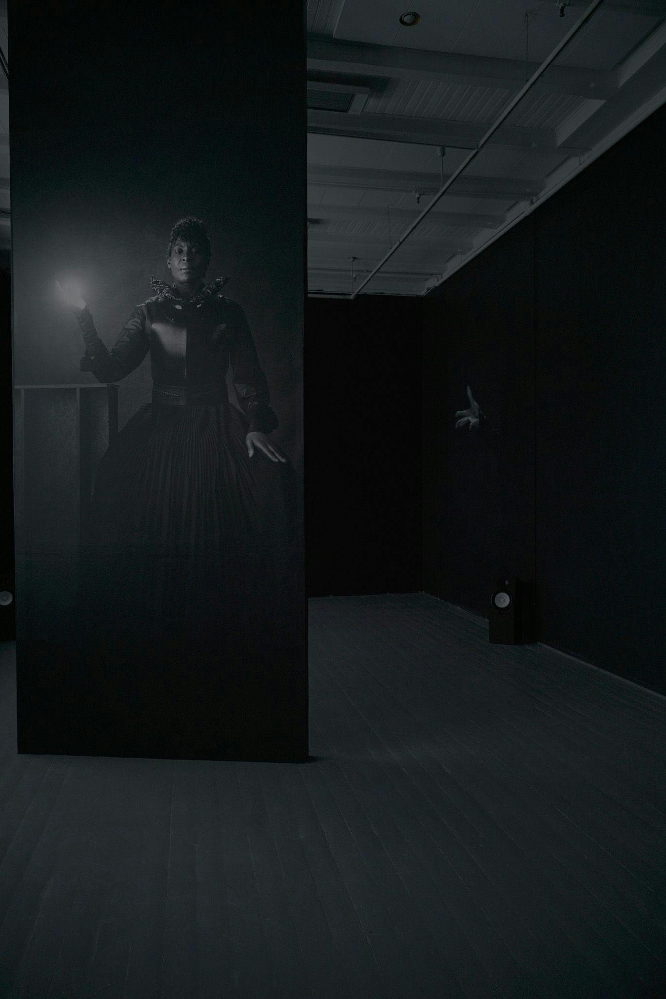En dame i en svart kjole som holderen lyskule blir projisert på en svart vegg, i et svart rom.