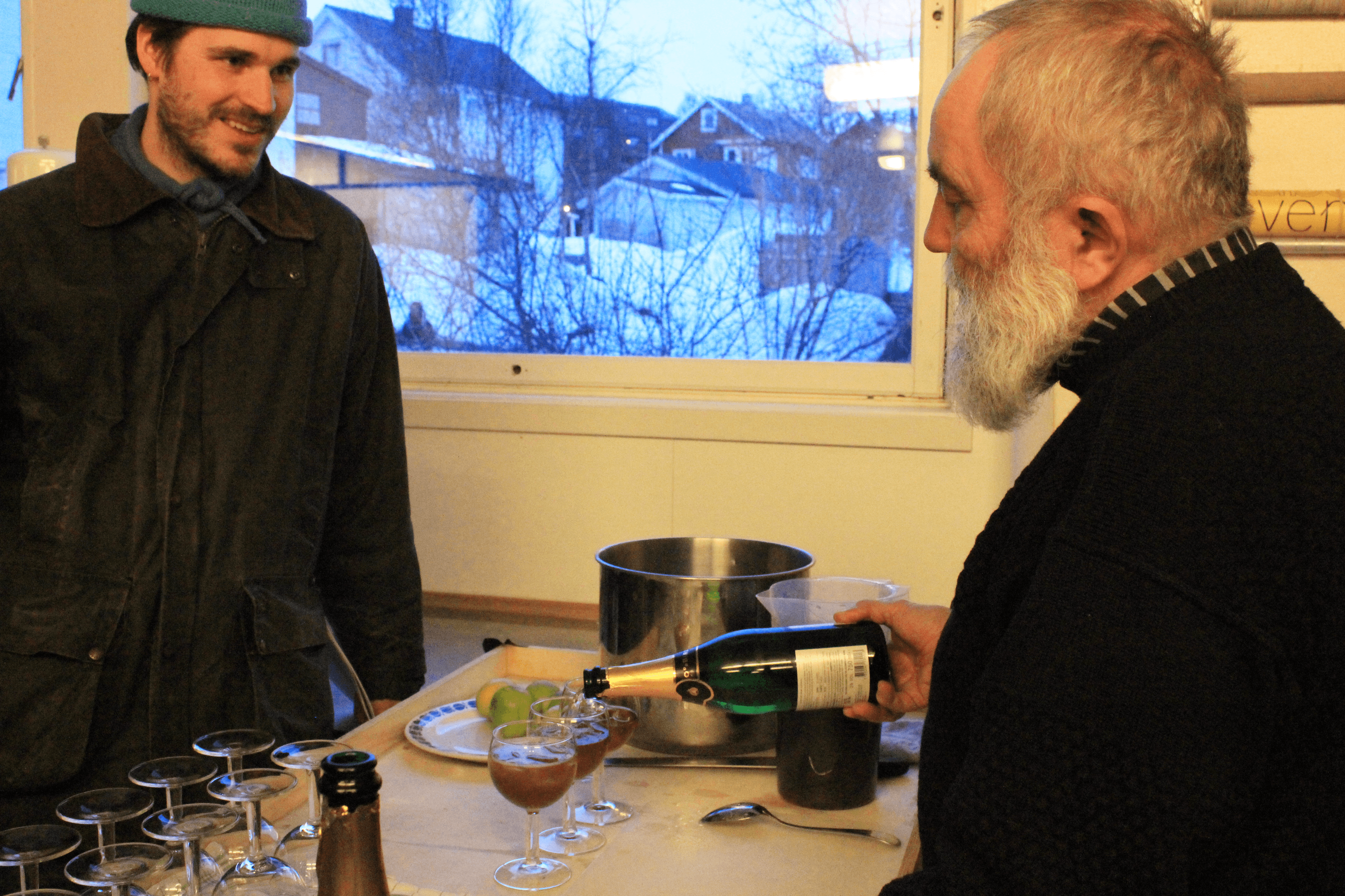 A man pours out a dark cocktail from a glass bottle. A man stands opposite smiling.