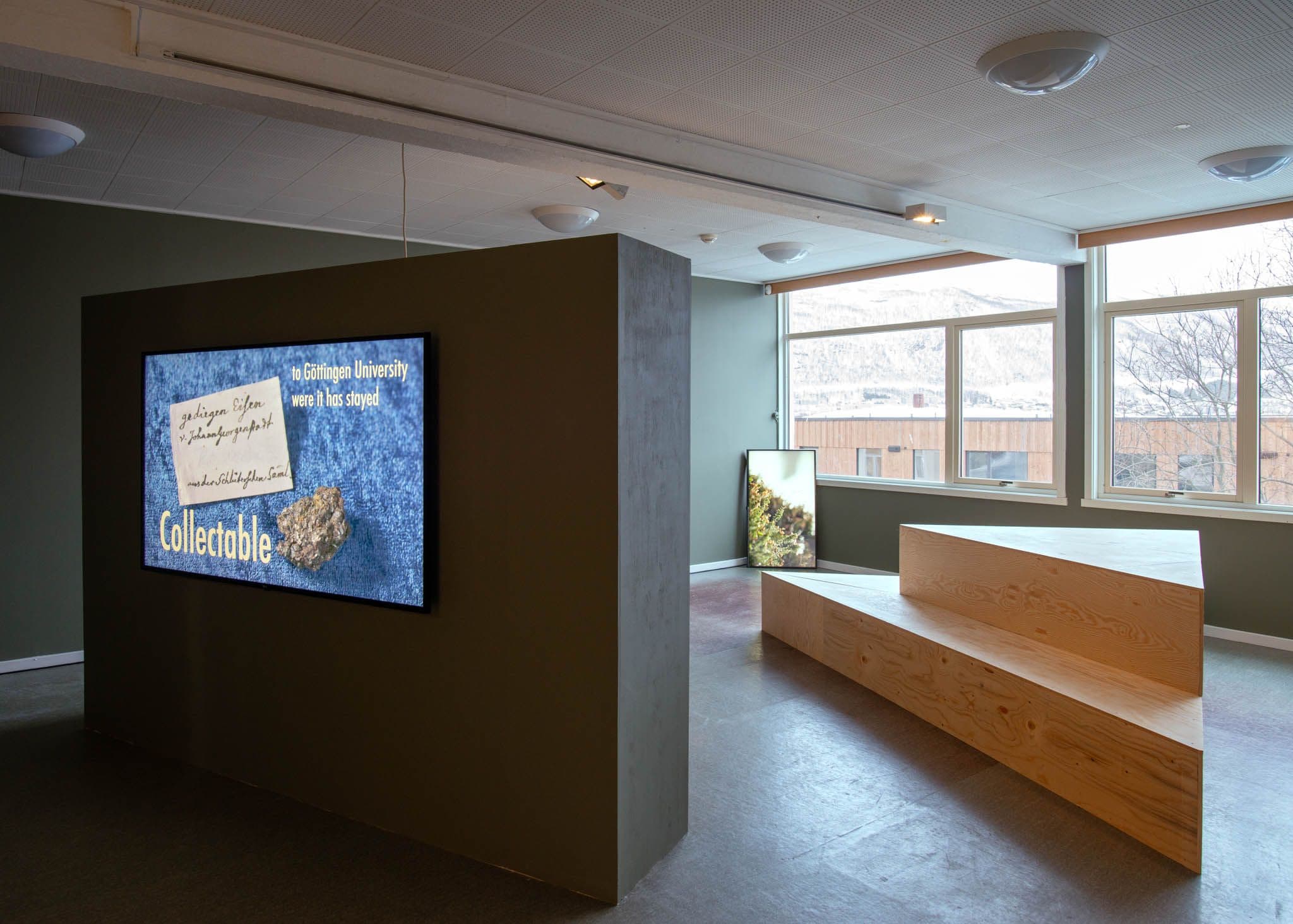 Large TV mounted to a freestanding wall in foreground. Behind is a triangular bench in front of windows, and a TV leaning vertically on the wall.