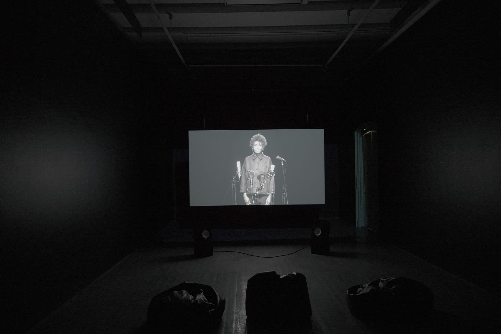 En projeksjonslerret vises en kvinne som står foran mikrofoner i svart-hvitt.