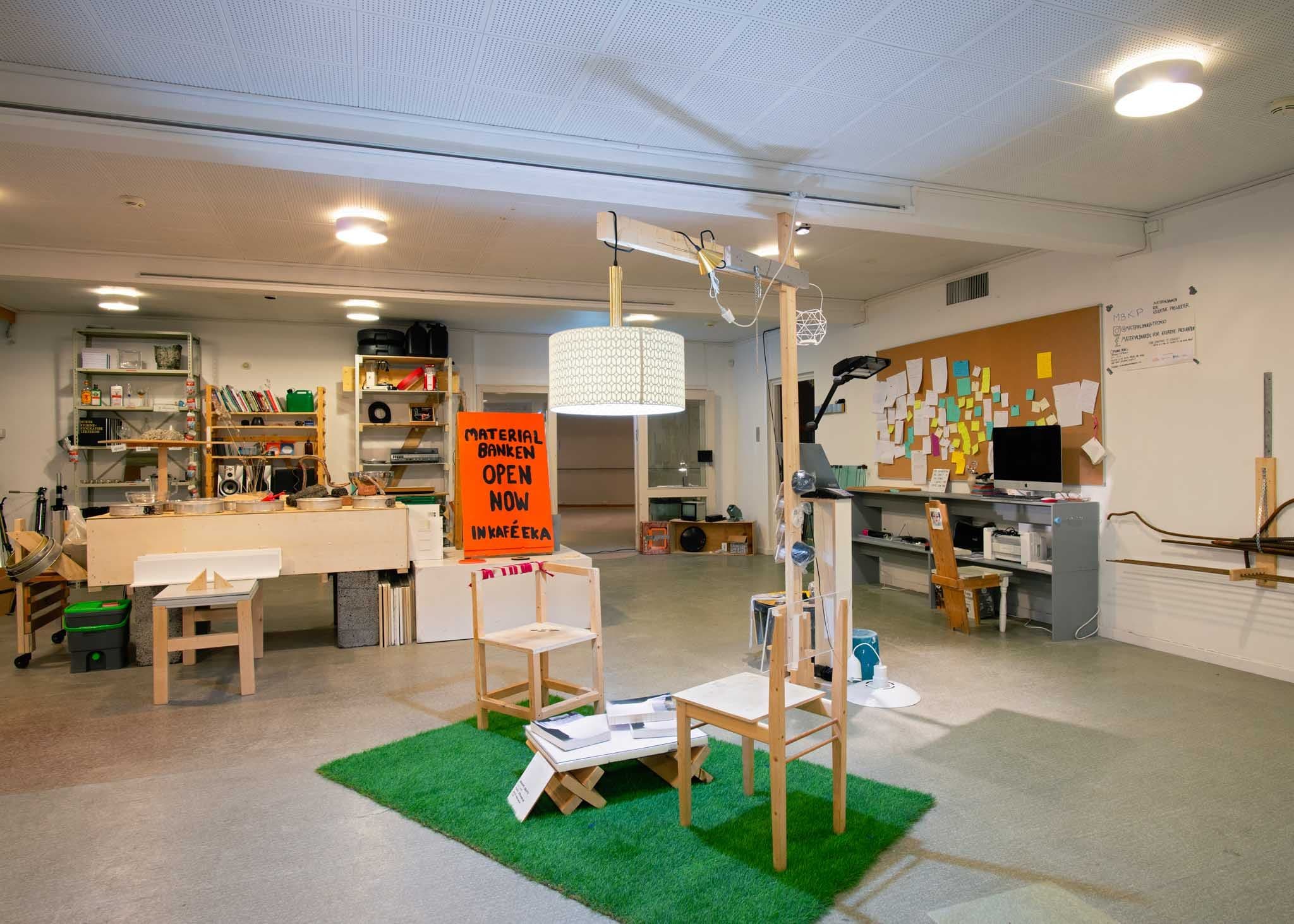 Two DIY chairs stand under a wooden lamp on artificial grass. Random objects are displayed in the background.